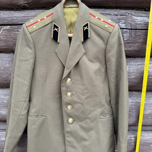 May include: An olive green military jacket with gold buttons and shoulder epaulets with red stripes and star details. The jacket has a notched collar and is suspended on a wooden hanger. The background is wooden. A vintage military garment.