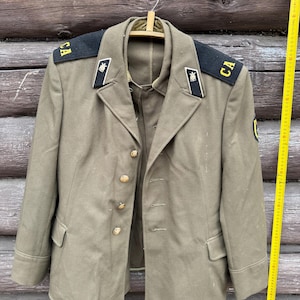 May include: An olive green military jacket with gold buttons and shoulder epaulets. The jacket has a notched collar, two front pockets, and is hanging on a wooden hanger. The epaulets have the letters "CA" on them. A yellow measuring tape is visible.
