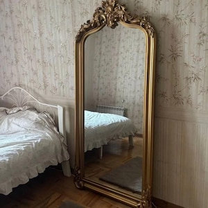 Peut inclure: Un grand miroir sur pied doté d'un cadre doré arqué et orné. Le cadre présente des détails sculptés complexes en haut et en bas, avec une surface en verre lisse et réfléchissante qui montre le reflet d'un intérieur de chambre.