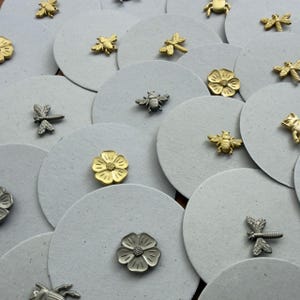 Puede incluir: Un surtido de pequeños adornos metálicos con forma de insectos y flores. La colección incluye libélulas, abejas y flores en tonos dorados y plateados, dispuestos sobre fondos circulares grises. Estos elementos decorativos son adecuados para manualidades y joyería.