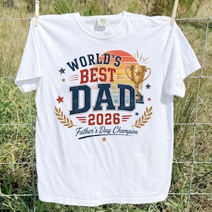 May include: White t-shirt with the text "WORLD'S BEST DAD 2026 Father's Day Champion" in blue, red, and gold. The shirt is hanging on a clothesline in front of a grassy field and a blue sky.