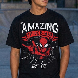 Puede incluir: Camiseta negra con un gr&aacute;fico de Spider-Man y el texto "THE AMAZING SPIDER-MAN" en blanco y rojo. El dise&ntilde;o incluye una telara&ntilde;a y el texto "Est. '62".