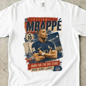 Puede incluir: Camiseta blanca con un dise&ntilde;o gr&aacute;fico de Kylian Mbapp&eacute;, futbolista, con el nombre "MBAPPE" en un lugar destacado. El dise&ntilde;o incluye el n&uacute;mero 10, la Torre Eiffel y la frase "BORN FOR THE BIG STAGE".