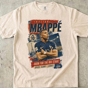 Puede incluir: Camiseta color marfil con un gr&aacute;fico de Kylian Mbapp&eacute;, futbolista. El dise&ntilde;o incluye el nombre del jugador, el n&uacute;mero 10 y el texto "BORN FOR THE BIG STAGE". La camiseta tambi&eacute;n tiene las palabras "SPEED GLORY LEGACY" y el a&ntilde;o 1998.