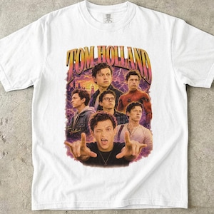 May include: White t-shirt featuring a graphic of Tom Holland in various poses, with the text "TOM HOLLAND" in a retro style. The design includes a purple and orange background.