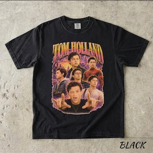 May include: Black t-shirt featuring a graphic of Tom Holland in various poses, with the name "TOM HOLLAND" in a retro font. The design includes images of the actor against a purple and orange background. The word "BLACK" is at the bottom.