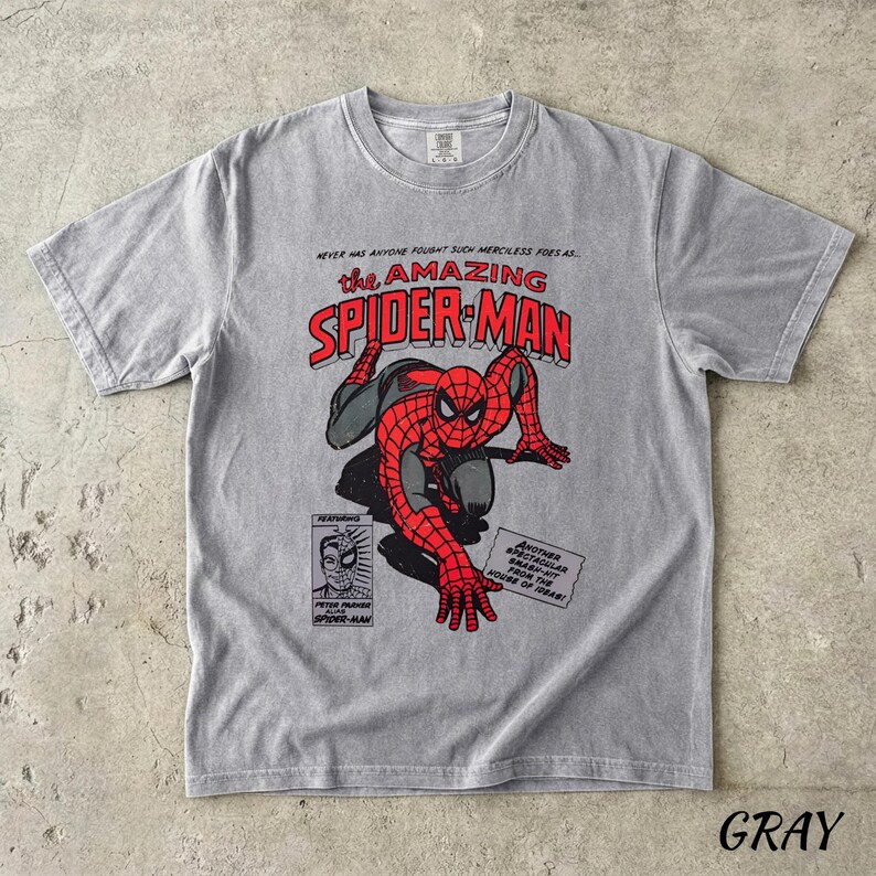 May include: Gray t-shirt featuring a comic book graphic of Spider-Man in red and black. The text "the AMAZING SPIDER-MAN" is in red and white. Additional text includes "NEVER HAS ANYONE FOUGHT SUCH MERCILESS FOES AS..." and "GRAY".