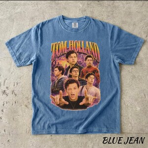 May include: Blue jean-colored t-shirt featuring a graphic of Tom Holland with the text "TOM HOLLAND" in a retro style. The shirt has a crew neck and short sleeves. The words "BLUE JEAN" are printed at the bottom.