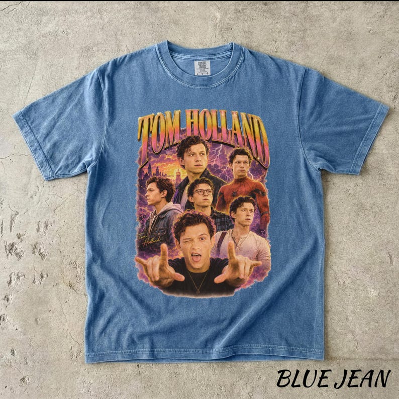 May include: Blue jean-colored t-shirt featuring a graphic of Tom Holland with the text "TOM HOLLAND" in a retro style. The shirt has a crew neck and short sleeves. The words "BLUE JEAN" are printed at the bottom.
