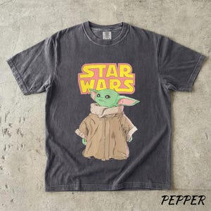 May include: Dark gray t-shirt featuring a graphic of Grogu from Star Wars, with the text "STAR WARS" in pink and yellow. The shirt has a crew neck and short sleeves. The word "PEPPER" is printed at the bottom.