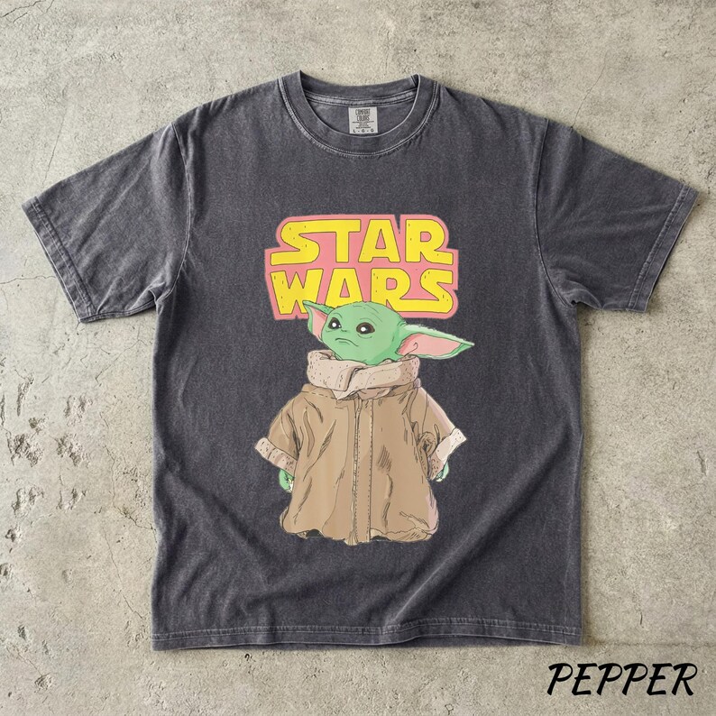 May include: Dark gray t-shirt featuring a graphic of Grogu from Star Wars, with the text "STAR WARS" in pink and yellow. The shirt has a crew neck and short sleeves. The word "PEPPER" is printed at the bottom.