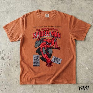 May include: A burnt orange t-shirt featuring a comic book style graphic of Spider-Man. The design includes the text "the AMAZING SPIDER-MAN" in red and white, with additional text and comic panels.
