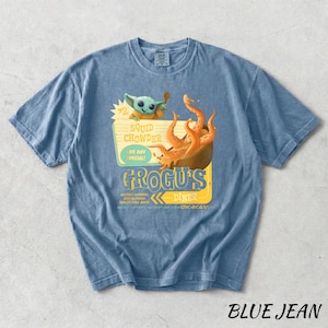 Puede incluir: Camiseta azul vaquero con un gr&aacute;fico de Grogu's Diner, con un personaje de dibujos animados y un cuenco de sopa de calamares. La camiseta tiene el texto "Squid Chowder, Life Day Special!" y "Grogus Diner".