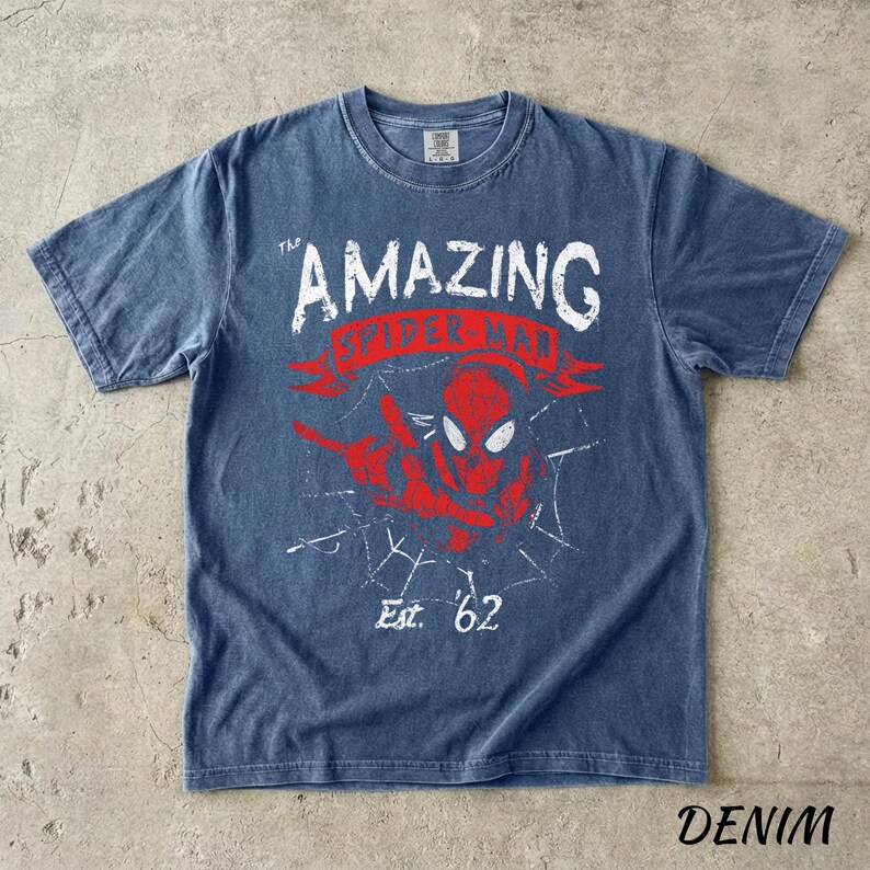 Puede incluir: Camiseta azul denim con un dise&ntilde;o desgastado de Spider-Man. El texto dice "THE AMAZING SPIDER-MAN" y "Est. '62". El dise&ntilde;o incluye una imagen roja de Spider-Man y una telara&ntilde;a.