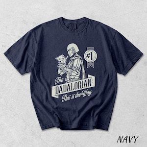 Pu&ograve; includere: T-shirt blu navy con grafica bianca di un personaggio di una popolare serie di fantascienza che tiene in braccio una piccola creatura. Il testo "The DADALORIAN This is the Way" &egrave; visualizzato sotto, con un distintivo "#1".