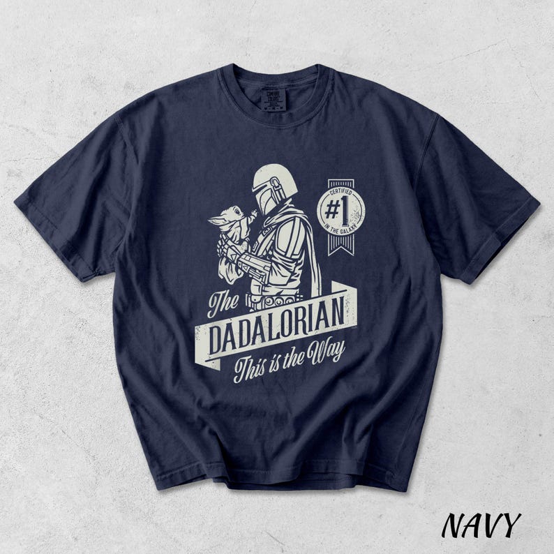 Pu&ograve; includere: T-shirt blu navy con grafica bianca di un personaggio di una popolare serie di fantascienza che tiene in braccio una piccola creatura. Il testo "The DADALORIAN This is the Way" &egrave; visualizzato sotto, con un distintivo "#1".