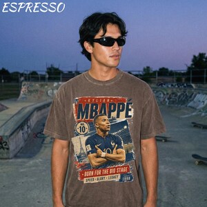 Puede incluir: Una camiseta marr&oacute;n con un gr&aacute;fico de Kylian Mbapp&eacute;, el texto "MBAPPE" en rojo y azul, y las palabras "BORN FOR THE BIG STAGE" y "SPEED GLORY LEGACY 1998". La palabra "ESPRESSO" est&aacute; en la parte superior.