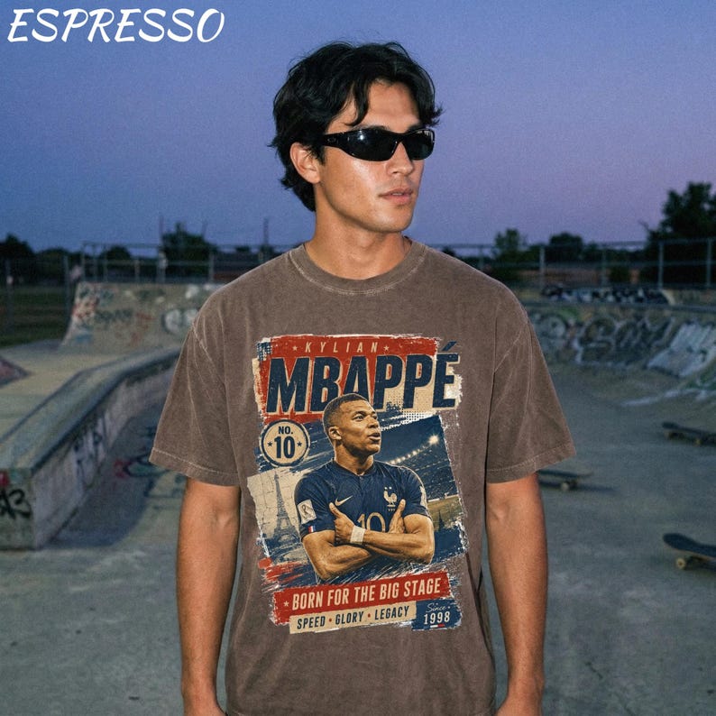 Puede incluir: Una camiseta marr&oacute;n con un gr&aacute;fico de Kylian Mbapp&eacute;, el texto "MBAPPE" en rojo y azul, y las palabras "BORN FOR THE BIG STAGE" y "SPEED GLORY LEGACY 1998". La palabra "ESPRESSO" est&aacute; en la parte superior.