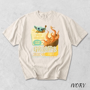 Puede incluir: Camiseta de color marfil con un dise&ntilde;o de dibujos animados. El gr&aacute;fico incluye un personaje alien&iacute;gena verde, un cuenco de tent&aacute;culos naranjas y el texto "Squid Chowder" y "Grogus Diner". La camiseta tiene cuello redondo y mangas cortas.