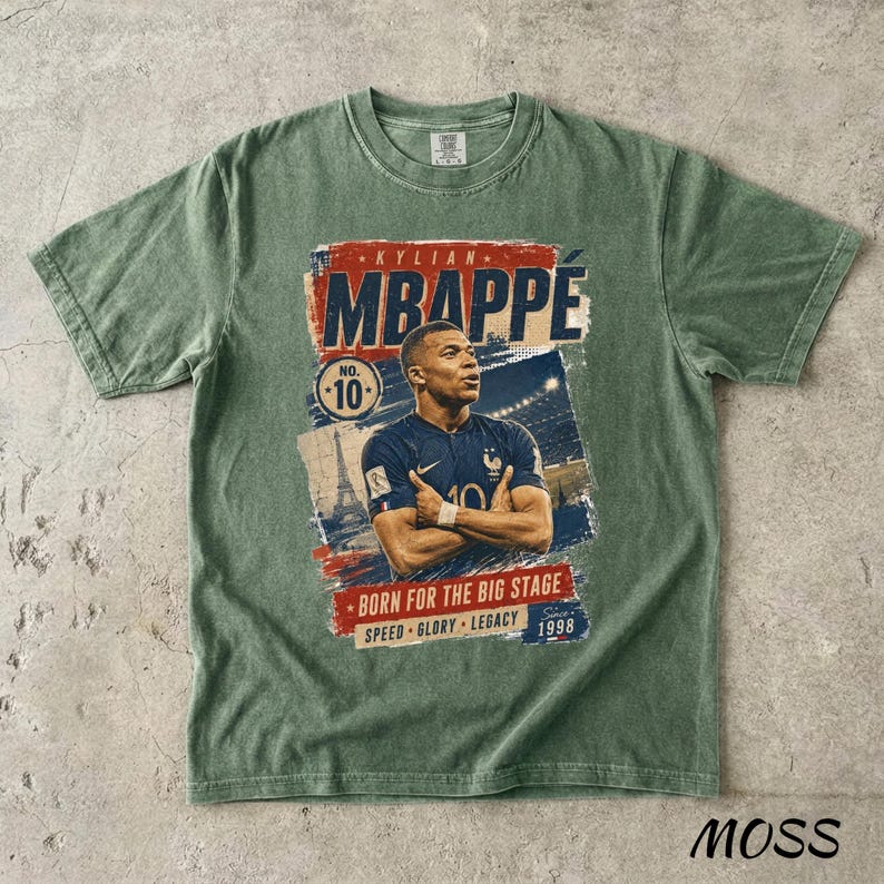 Puede incluir: Camiseta verde musgo con un gr&aacute;fico de Kylian Mbapp&eacute;. El dise&ntilde;o incluye el texto "MBAPPE" en rojo y azul, junto con "BORN FOR THE BIG STAGE" y "1998". Tambi&eacute;n aparece el n&uacute;mero 10. La camiseta est&aacute; hecha de un material suave.