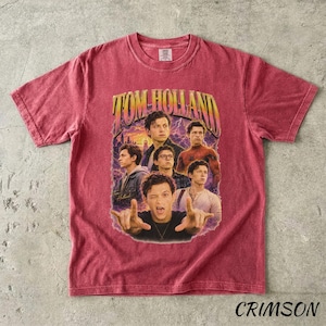 May include: Crimson-colored t-shirt featuring a vintage-style graphic of Tom Holland. The graphic includes multiple images of the actor with the text "TOM HOLLAND" in a retro font. The shirt is a crew neck.