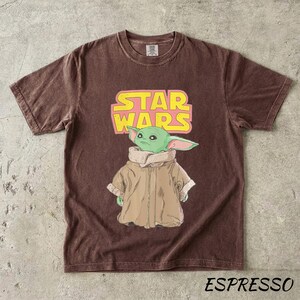 May include: Espresso-colored t-shirt featuring a graphic of a green alien character from Star Wars. The Star Wars logo is in yellow and pink. The shirt is a crew neck and has short sleeves.