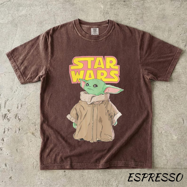 May include: Espresso-colored t-shirt featuring a graphic of a green alien character from Star Wars. The Star Wars logo is in yellow and pink. The shirt is a crew neck and has short sleeves.