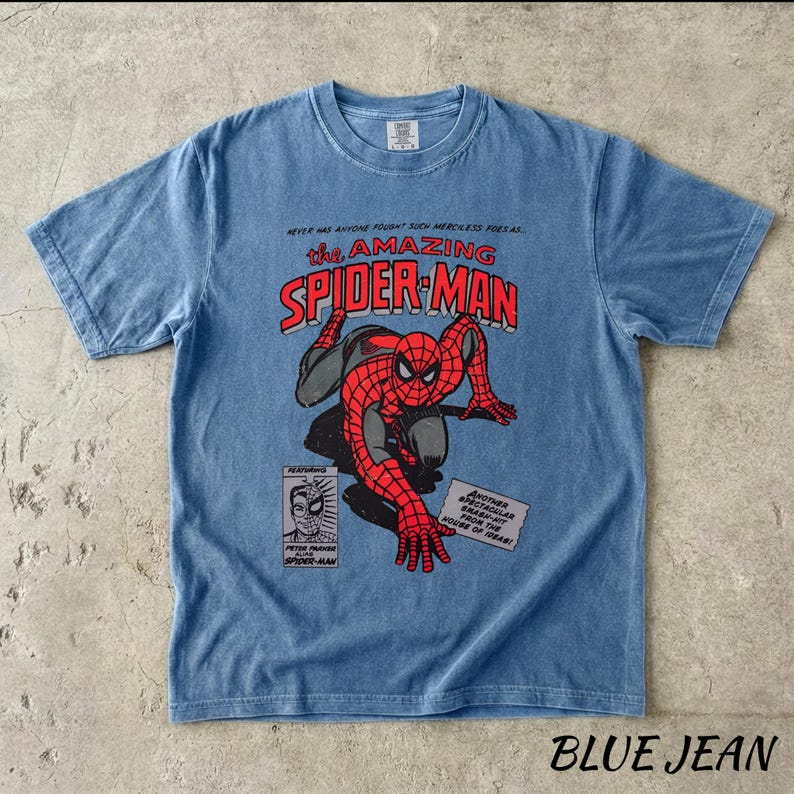May include: Blue jean colored t-shirt featuring a vintage comic book style graphic of Spider-Man. The design includes the text "the AMAZING SPIDER-MAN" in red and white, with additional comic book panels and text.