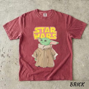 May include: Brick-colored t-shirt featuring a graphic of a green alien character from Star Wars. The Star Wars logo is in yellow and pink. The word "BRICK" is printed at the bottom of the shirt.