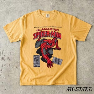 May include: Mustard-colored t-shirt featuring a comic book graphic of Spider-Man. The design includes the text "The Amazing Spider-Man" in red and white, with a drawing of Spider-Man in his classic red and black suit. Additional text is visible.