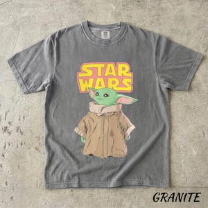 May include: Gray t-shirt featuring a graphic of a green alien character from Star Wars, with the words "STAR WARS" in yellow and pink. The character is wearing a brown coat. The word "GRANITE" is printed at the bottom of the shirt.