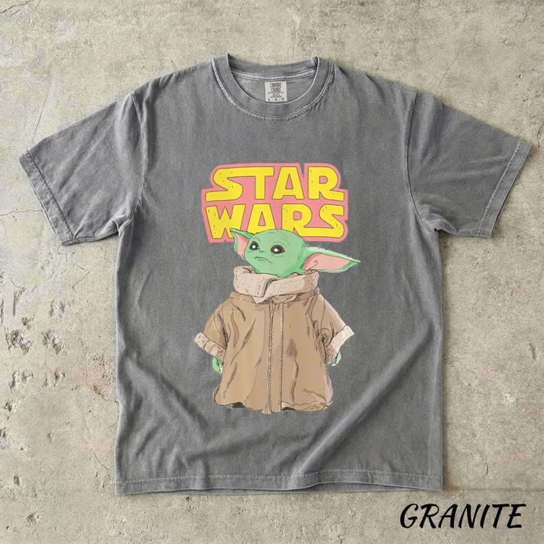 May include: Gray t-shirt featuring a graphic of a green alien character from Star Wars, with the words "STAR WARS" in yellow and pink. The character is wearing a brown coat. The word "GRANITE" is printed at the bottom of the shirt.