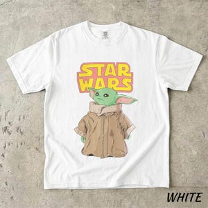 May include: White t-shirt featuring a graphic of a green alien character from Star Wars, with the text "STAR WARS" in yellow and pink. The alien is wearing a brown coat. The word "WHITE" is printed at the bottom.
