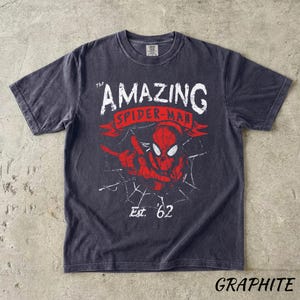 Puede incluir: Camiseta de color grafito con un dise&ntilde;o desgastado de Spider-Man, con el texto "The Amazing Spider-Man" y "Est. '62". El dise&ntilde;o incluye una imagen roja de Spider-Man y detalles de telara&ntilde;a.