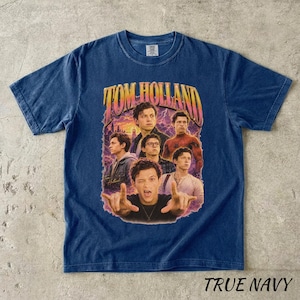 May include: A navy blue t-shirt featuring a graphic of Tom Holland with the text "TOM HOLLAND" in a retro style. The shirt has a crew neck and short sleeves. The words "TRUE NAVY" are printed at the bottom.