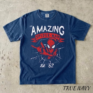 Puede incluir: Camiseta azul marino con un gr&aacute;fico de Spider-Man, el texto "The Amazing Spider-Man" y "Est. '62". El dise&ntilde;o es rojo y blanco. La camiseta est&aacute; etiquetada como "True Navy".