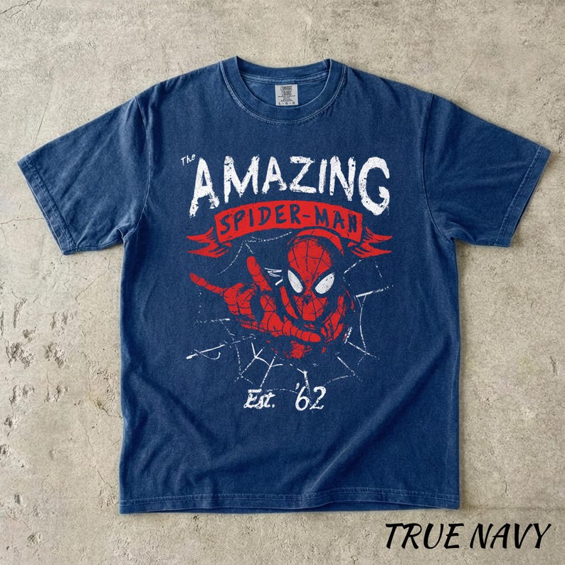 Puede incluir: Camiseta azul marino con un gr&aacute;fico de Spider-Man, el texto "The Amazing Spider-Man" y "Est. '62". El dise&ntilde;o es rojo y blanco. La camiseta est&aacute; etiquetada como "True Navy".