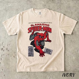 May include: Ivory-colored t-shirt featuring a comic book graphic of Spider-Man in red and black. The text "the AMAZING SPIDER-MAN" is displayed above the character. Additional text includes "NEVER HAS ANYONE FOUGHT SUCH MERCILESS FOES AS..." and "FEATURING".