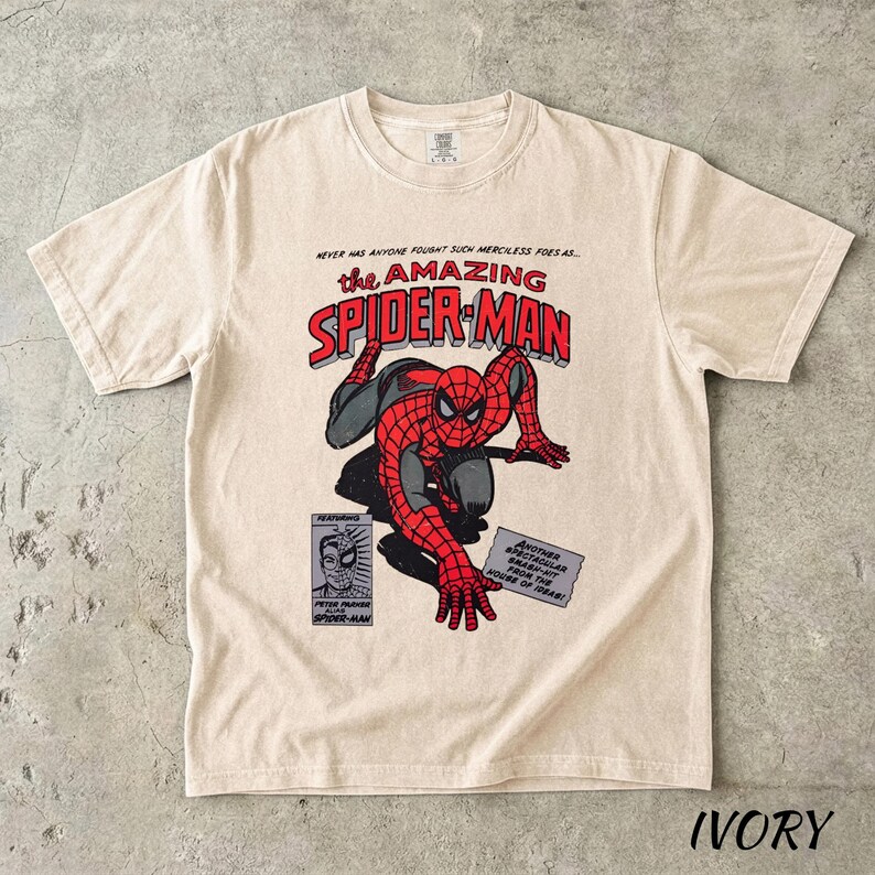 May include: Ivory-colored t-shirt featuring a comic book graphic of Spider-Man in red and black. The text "the AMAZING SPIDER-MAN" is displayed above the character. Additional text includes "NEVER HAS ANYONE FOUGHT SUCH MERCILESS FOES AS..." and "FEATURING".