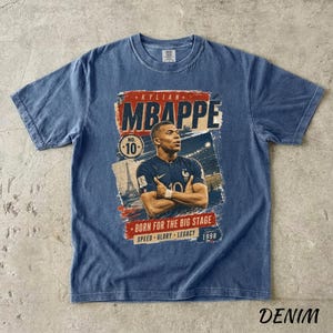Puede incluir: Camiseta azul denim con un gr&aacute;fico de Kylian Mbapp&eacute;, que muestra el texto "MBAPPE" en rojo y blanco. La camiseta incluye el n&uacute;mero 10 y las frases "BORN FOR THE BIG STAGE", "SPEED GLORY LEGACY" y el a&ntilde;o "1998".