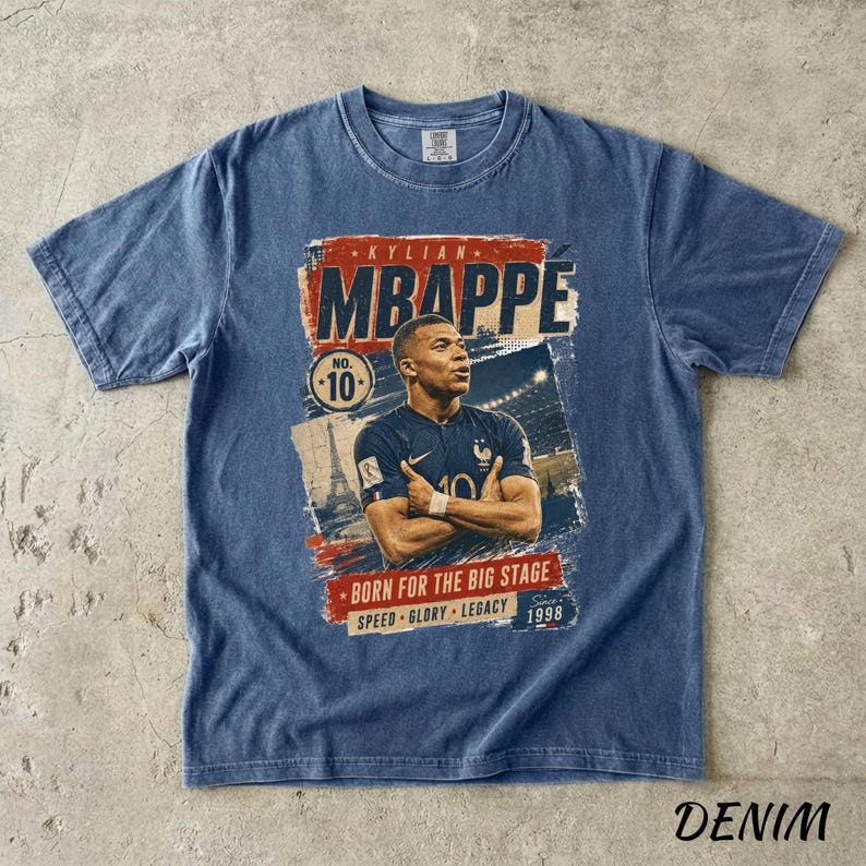 Puede incluir: Camiseta azul denim con un gr&aacute;fico de Kylian Mbapp&eacute;, que muestra el texto "MBAPPE" en rojo y blanco. La camiseta incluye el n&uacute;mero 10 y las frases "BORN FOR THE BIG STAGE", "SPEED GLORY LEGACY" y el a&ntilde;o "1998".