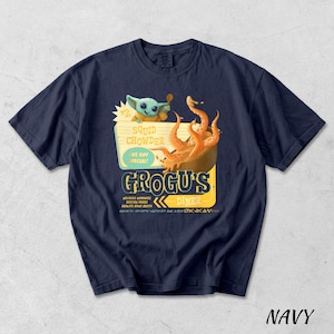 Puede incluir: Camiseta azul marino con un dise&ntilde;o de dibujos animados. El dise&ntilde;o incluye un personaje alien&iacute;gena verde y un cuenco de tent&aacute;culos naranjas, con el texto "Squid Chowder" y "Grogu's Diner". La palabra "Navy" est&aacute; en la parte inferior.