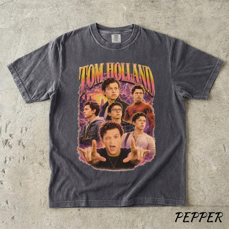 May include: Dark gray t-shirt featuring a vintage-style graphic of Tom Holland in various poses. The graphic includes the text "TOM HOLLAND" in a retro font, with a purple and orange color scheme. The shirt has a classic crew neck and short sleeves.