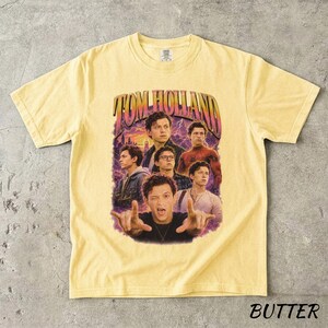 May include: Butter-colored t-shirt featuring a graphic of Tom Holland with the text "TOM HOLLAND" in a retro design. The shirt showcases multiple images of the actor against a purple and blue background.