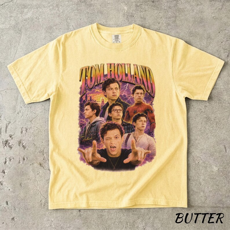 May include: Butter-colored t-shirt featuring a graphic of Tom Holland with the text "TOM HOLLAND" in a retro design. The shirt showcases multiple images of the actor against a purple and blue background.