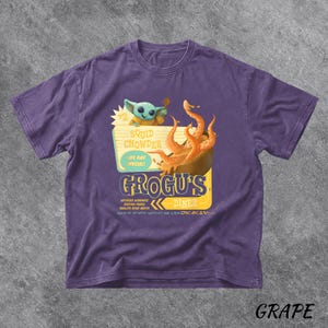 Puede incluir: Camiseta morada con un dise&ntilde;o de Grogu's Diner, con el texto "Squid Chowder" y "Life Day Special!". El dise&ntilde;o incluye una ilustraci&oacute;n de Grogu y un cuenco de comida. La palabra "Grape" est&aacute; en la parte inferior.