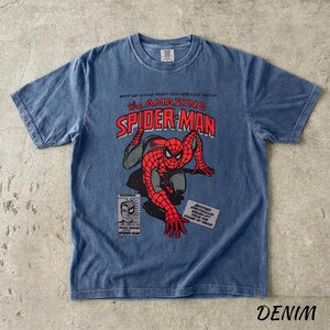 May include: Denim blue t-shirt featuring a graphic of Spider-Man from the comic book series "The Amazing Spider-Man." The design includes the title, a drawing of Spider-Man, and comic book panels. The shirt has a crew neck and short sleeves.
