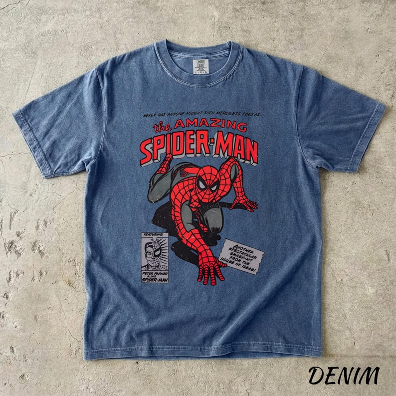 May include: Denim blue t-shirt featuring a graphic of Spider-Man from the comic book series "The Amazing Spider-Man." The design includes the title, a drawing of Spider-Man, and comic book panels. The shirt has a crew neck and short sleeves.