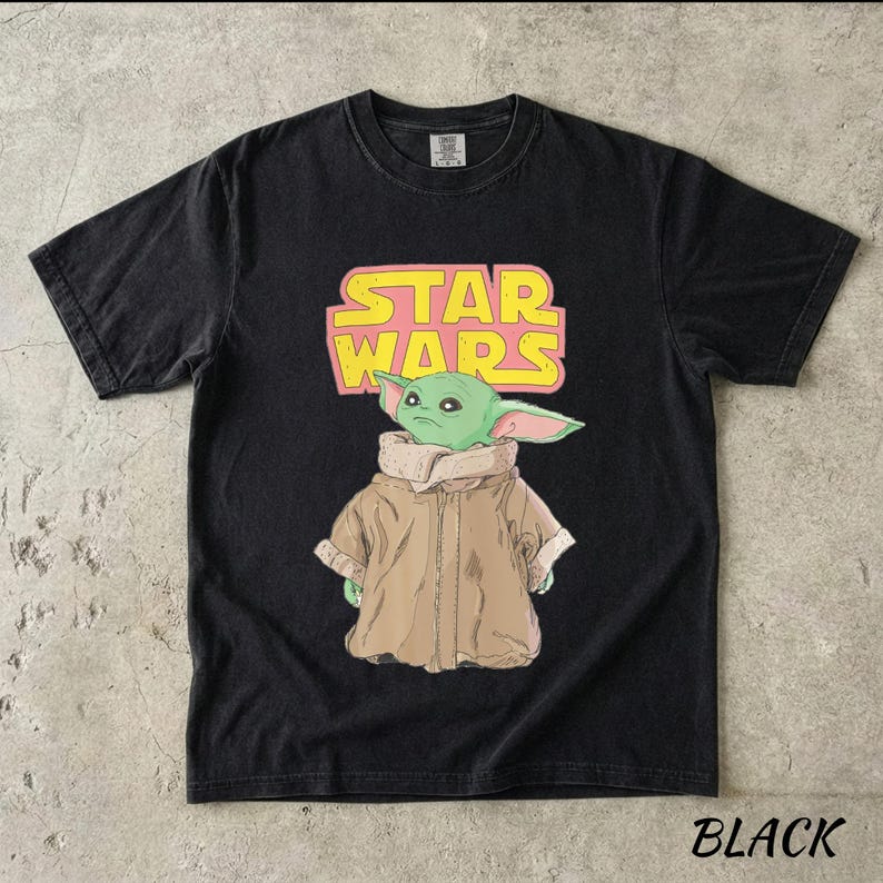 May include: Black t-shirt featuring a graphic of a green alien character from Star Wars, with the text "STAR WARS" in pink and yellow. The word "BLACK" is printed at the bottom. The shirt has short sleeves and a crew neck.