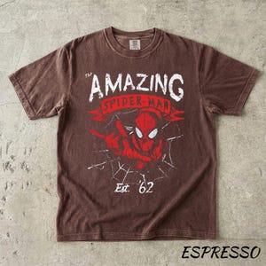 Puede incluir: Camiseta color espresso con un gr&aacute;fico desgastado de Spider-Man. El dise&ntilde;o incluye el texto "THE AMAZING SPIDER-MAN" en blanco y rojo, una telara&ntilde;a y "Est. '62".